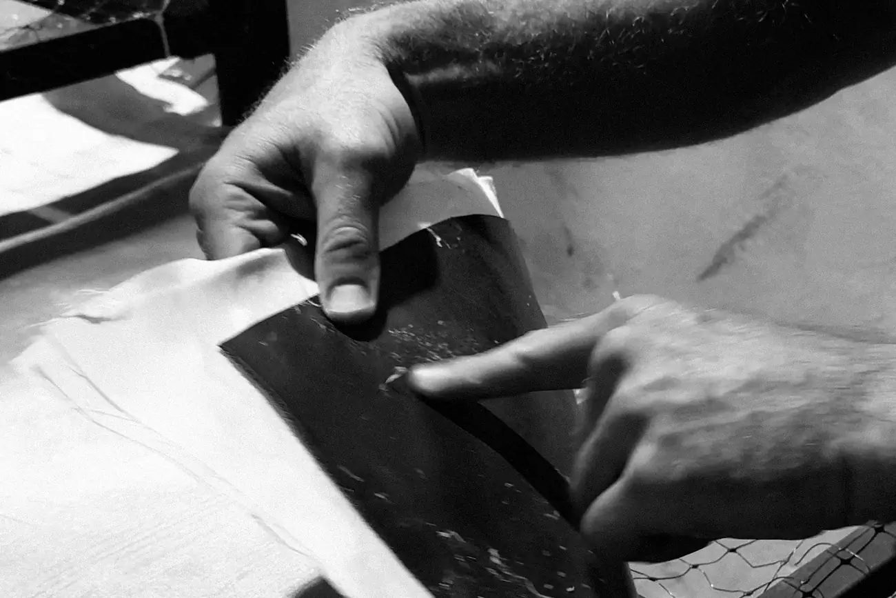 Two hands peeling a layer back from a latex-coated canvas sheet in the workshop, in black and white.