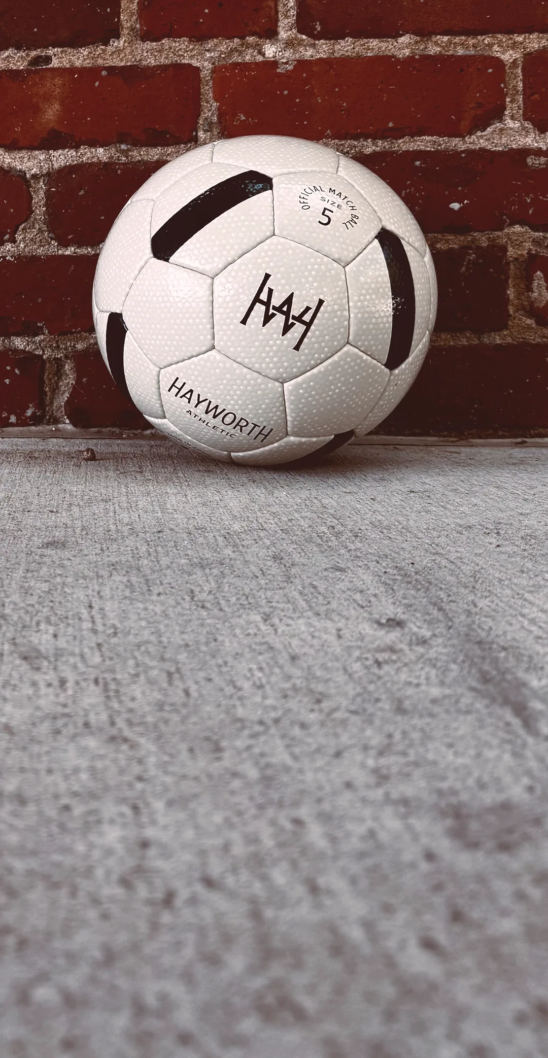 A Hayworth Athletic Standard Model No. 152 soccer ball resting on a concrete sidewalk against a weathered red brick wall.