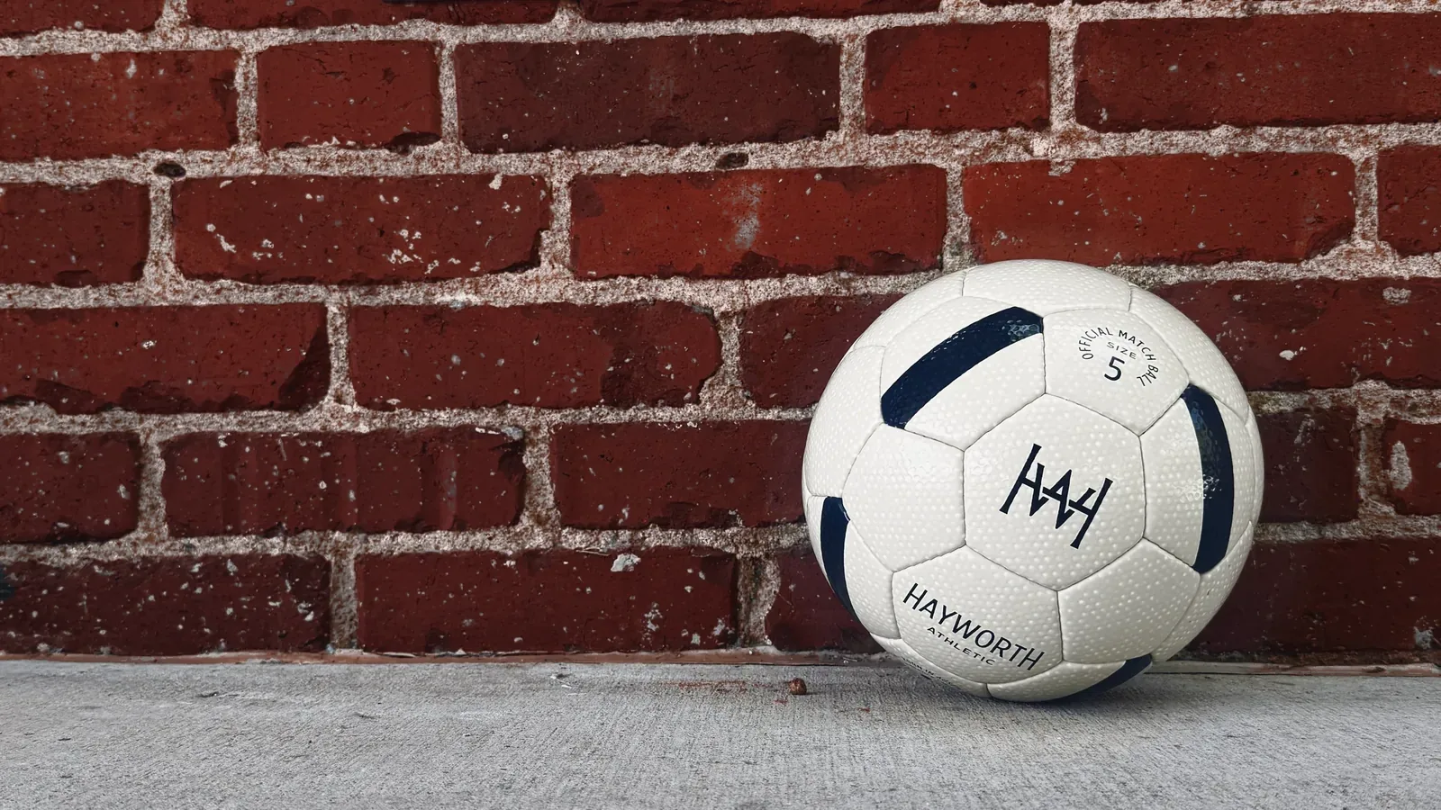 A Hayworth Athletic Standard Model No. 152 soccer ball resting on a concrete sidewalk against a weathered red brick wall.